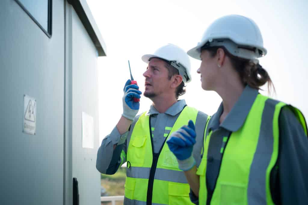 A team of electrical engineers is inspecting an energy storage station of a solar panels in the middle of a hundred acre field of solar panels.