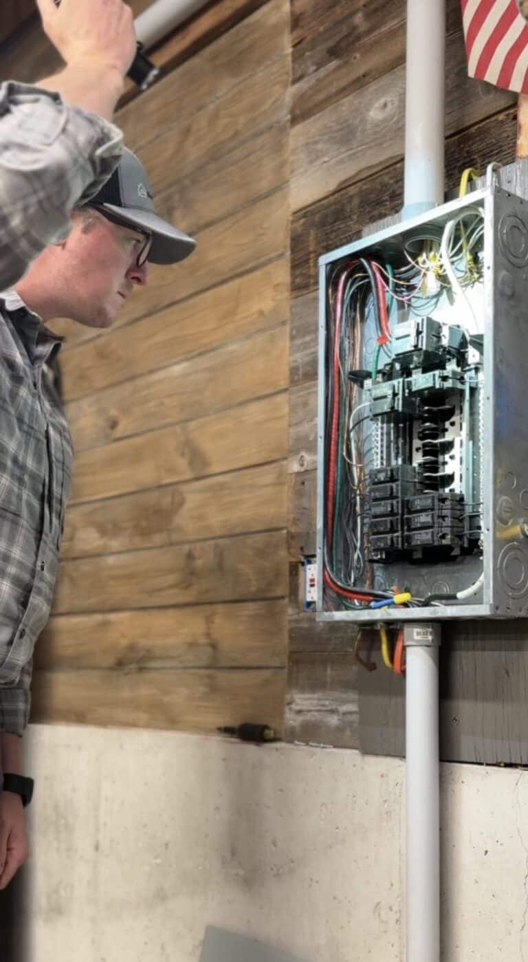 Home inspector examining an open electrical panel on a wooden wall, using a flashlight to check wiring and circuit breakers during a residential inspection.